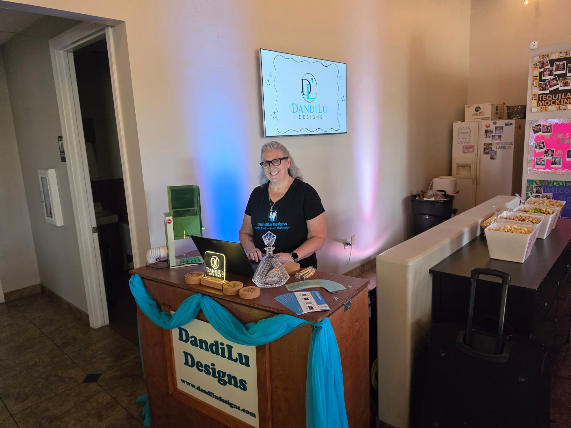 Randi behind the DandiLu Designs engraving bar at an indoor event with branded signage and teal draping